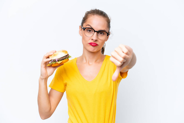 Mujer caucásica joven sosteniendo una hamburguesa aislada sobre fondo blanco mostrando el pulgar hacia abajo con expresión negativa - Foto, imagen