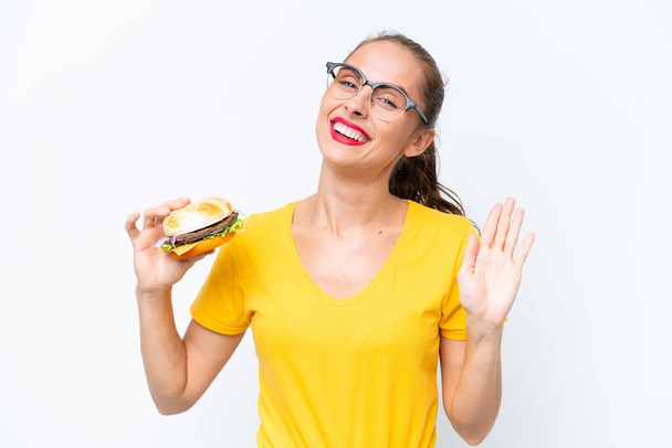 Young caucasian woman holding a burger isolated on white background saluting with hand with happy expression - Fotó, kép