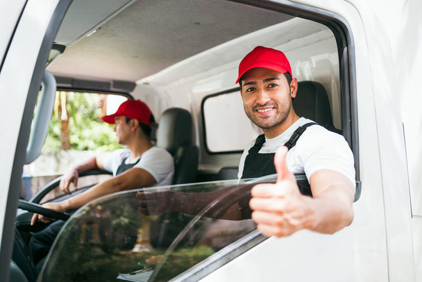 Boldog hivatásos kamionsofőr, akinek az asszisztense piros sapkát visel, mosolyog, belenéz a teherautó ablakából a kamerába, mielőtt kiszállítja a csomagot. A teherautó-vezető és a szállítási szolgáltatás koncepciója. - Fotó, kép