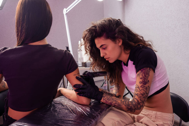 Επαγγελματική γυναίκα τατουάζ με εστιασμένη προσοχή επιδέξια τη δημιουργία εξατομικευμένων επιστολόχαρτο σχεδιασμό στο βραχίονα του πελάτη, χρησιμοποιώντας μηχανή τατουάζ - Φωτογραφία, εικόνα