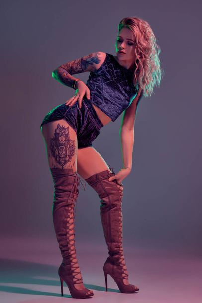 Nuori kaunis tatuoitu blondi nainen yllään sininen velour booty shortsit ja t-lyhyt, punaruskea mokka saappaat osoittaa hänen hämmästyttävä elin studiossa. - Valokuva, kuva