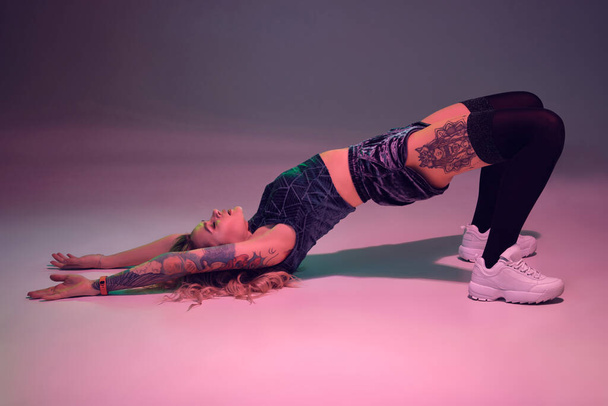 Young beautiful caucasian blonde girl with tattoo on her body, wearing velour blue booty shorts and black stockings making stretching exercises before dancing twerk, pink and blue background - Photo, Image