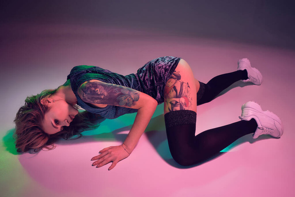 Joven hermosa chica rubia caucásica con tatuaje en su cuerpo, usando pantalones cortos de terciopelo azul botín y medias negras bailando twerk acostado con botín levantado, fondo rosa
 - Foto, imagen