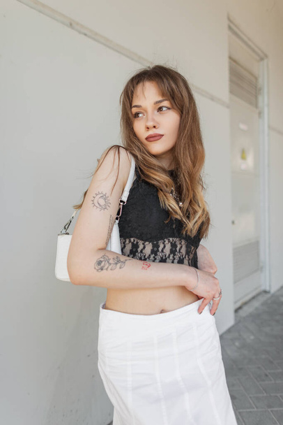 Moda bela mulher hipster com uma tatuagem no braço em um top de renda preta com um sutiã e uma saia branca com uma bolsa de mão caminha na cidade. Bonita senhora urbana - Foto, Imagem