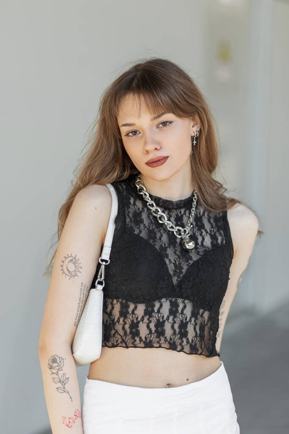 Όμορφη μόδα σέξι hipster γυναίκα μοντέλο με ένα τατουάζ στα χέρια της σε ένα μοντέρνο δαντέλα κορυφή με ένα μαύρο σουτιέν και μια λευκή φούστα με μια τσάντα βόλτες στην πόλη κοντά σε ένα γκρι κτίριο την ηλιόλουστη μέρα - Φωτογραφία, εικόνα