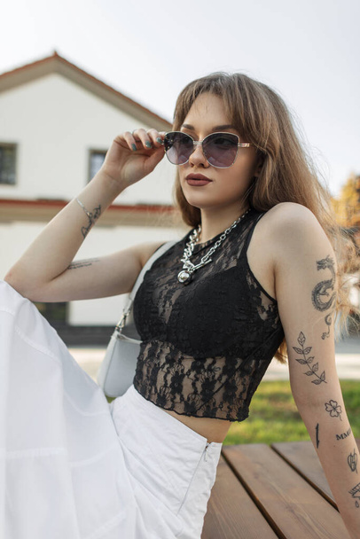 Chica hipster hermosa de moda en ropa de verano urbana con estilo con un top de encaje, sujetador negro y una falda con un bolso se sienta en un banco de la ciudad y se pone gafas de sol - Foto, imagen