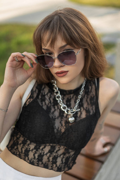 Fashionable young beautiful woman in fashion summer outfit with sexy black lace wears a stylish sunglasses and sits on the street - Photo, Image
