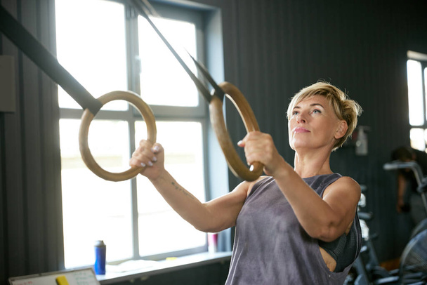 Mature woman training in gym, doing exercises with gymnastics rings. Hand pull-ups exercises. Strength. Concept of active, sportive and healthy lifestyle, fitness, body care, ad - Photo, Image