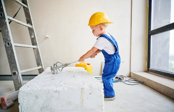 Pełna długość małego chłopca budowlańca za pomocą instrumentu budowlanego podczas pracy nad remontem domu. Dziecko w kombinezonach roboczych i kasku ochronnym przygotowujące materiał do prac remontowo-remontowych. - Zdjęcie, obraz