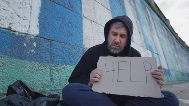 senzatetto povero giovane seduto a terra in città con un tavolo di aiuto mentre chiede soldi per sopravvivere. depressione, problema, sporco, sociale, tristezza, da solo, disperazione, strada. 4k di alta qualità - Filmati, video