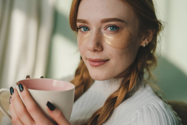 Retrato de mujer atractiva sonriente mirando a la cámara con parches para los ojos sosteniendo una taza de café en la sala de estar. Gente estilo de vida retrato. Residentes - Foto, Imagen