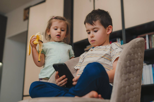 Dvě malé generace Z děti pomocí mobilního telefonu, hraní videohry pro děti, relaxaci na pohodlných křeslech. Digitální technologie. - Fotografie, Obrázek