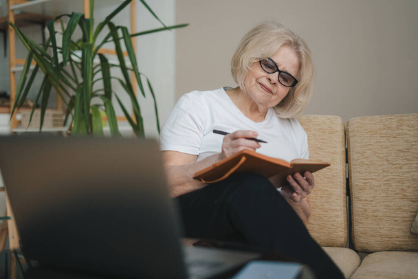 Atrakcyjna, pozytywna starsza kobieta siedząca przed laptopem i pisząca nową książkę. Biznesowy sukces. Nowoczesny styl życia. Ludzie korzystający z laptopa. - Zdjęcie, obraz