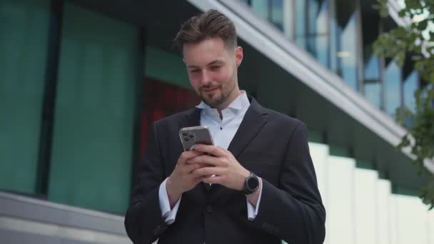 Tyylikäs komea liikemies tilalla puhelimen lukeminen sähköposti seisoo kadulla hymyillen kirjoittaa viestin kommunikoi sosiaalisen verkoston mietteliäs mies selaa tavaroita verkkokaupassa mobiilisovelluksella älypuhelimella - Materiaali, video