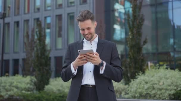 Elegante hombre de negocios guapo sosteniendo el correo electrónico de lectura de teléfono se para en la calle sonriendo escribe mensaje se comunica en la red social hombre pensativo navega mercancías en la tienda en línea utilizando la aplicación móvil en el teléfono inteligente - Imágenes, Vídeo