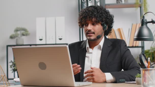 Індійський бізнесмен, який працює над ноутбуком, носячи навушники, колл-центр фрілансерів або оператор підтримки helpline, приємно спілкуючись з клієнтом або колегою по телефону в офісі - Кадри, відео