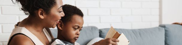 Black pleased mother and son reading book while sitting on sofa at home - Foto, immagini