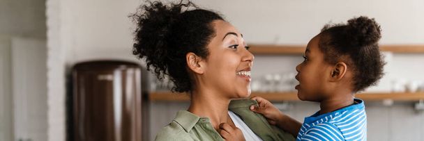Black happy mother laughing while holding her daughter at home kitchen - Photo, image