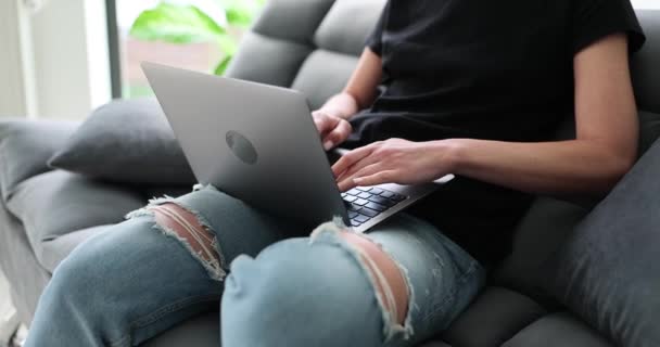 Female in ripped jeans sitting on couch in living room operates laptop on lap. Modern concept of device-based communication and remote working slow motion - Footage, Video