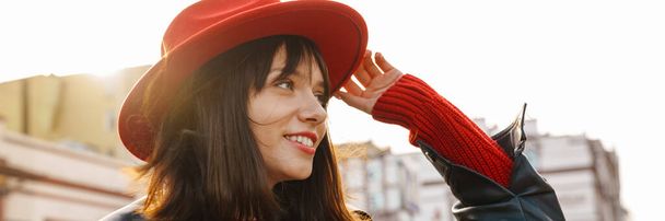 Close up of a smiling young stylish brunette white woman wearing hat and leather jacket with short hair standing on a street looking aside - Foto, afbeelding