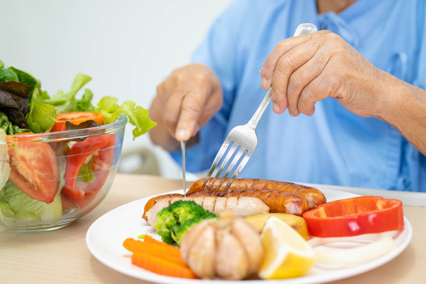 Asian senior woman patient eating Salmon steak breakfast with vegetable healthy food while sitting and hungry on bed in hospital. - Foto, immagini