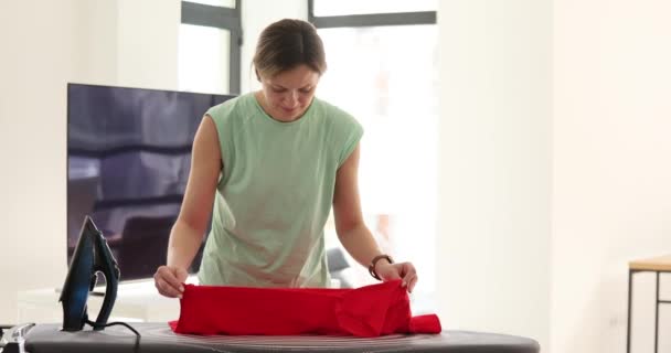 Ama de casa coloca camiseta roja en la tabla de planchar en la habitación luminosa del apartamento. Concepto de cuidado y habilidad en ropa y limpieza cámara lenta - Metraje, vídeo