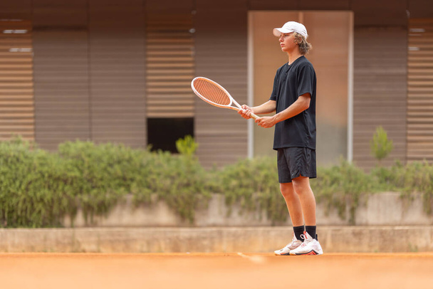 A boy in black t-shirt standing on tennis court holding a raquet. Mid shot - Photo, Image