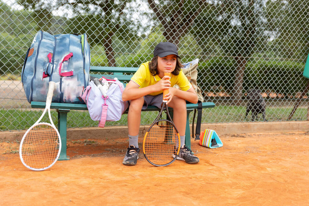 Een jongen zit op een bank met sportbafs bij de tennisbaan en wacht op zijn beurt. Tussenschot - Foto, afbeelding
