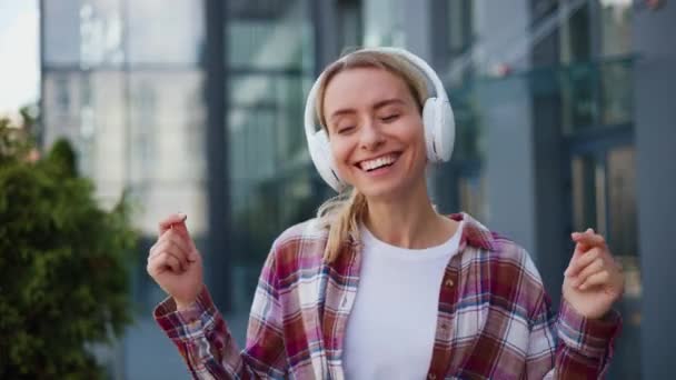 笑顔の若いポジティブな女性ヒップスターが街の外に立っている音楽を聴く - 映像、動画