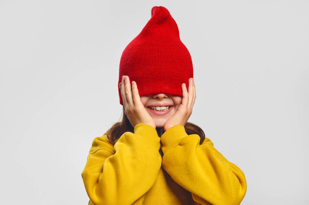 Entzückendes Hipster-Mädchen in gelbem Kapuzenpulli, das Augen mit rotem Hut bedeckt und auf weißem Hintergrund in die Kamera lächelt - Foto, Bild