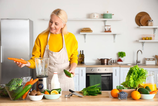 Ώριμη γυναίκα με φρέσκα λαχανικά και φρούτα κάνοντας υγιεινό smoothie στην κουζίνα - Φωτογραφία, εικόνα