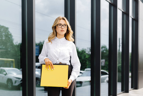 Элегантная, уверенная в себе молодая женщина, формально одетая, стоит на улице на фоне бизнес-центра, держа папку с документами, смотрит в камеру. - Фото, изображение