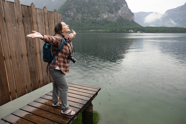 Giovane donna con una macchina fotografica in piedi su un palco di legno con le braccia aperte e sorridenti, Hallstatt, Austria. Foto di alta qualità - Foto, immagini