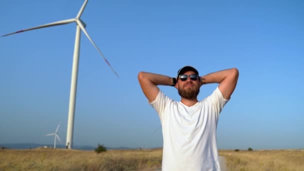 Feliz hombre barbudo relajado levantando brazos disfrutando de viajes en la naturaleza puesta de sol en el molino de viento de campo. Libertad, energía ecológica, consumo sostenible - Metraje, vídeo