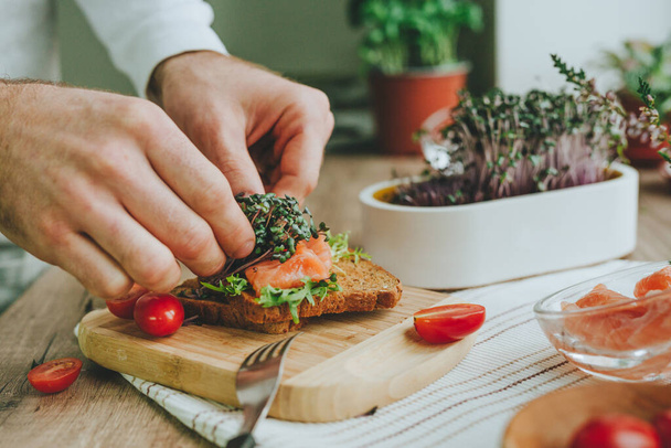 Close-up van mannetjes handen maken sandwich met zalm vis, radijs spruit microgroen en tomaat op houten achtergrond. Selectieve focus. - Foto, afbeelding