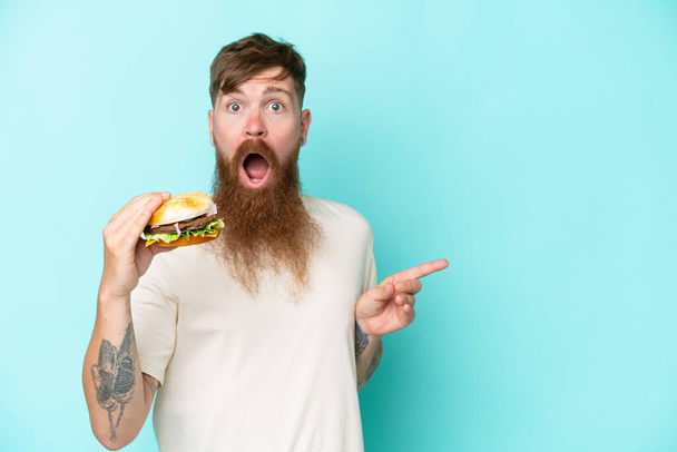 Vöröshajú férfi hosszú szakállal kezében egy burger elszigetelt kék háttér meglepett és ujjal mutogatott oldalra - Fotó, kép