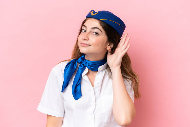Airplane stewardess caucasian woman isolated on pink background listening to something by putting hand on the ear - Photo, Image