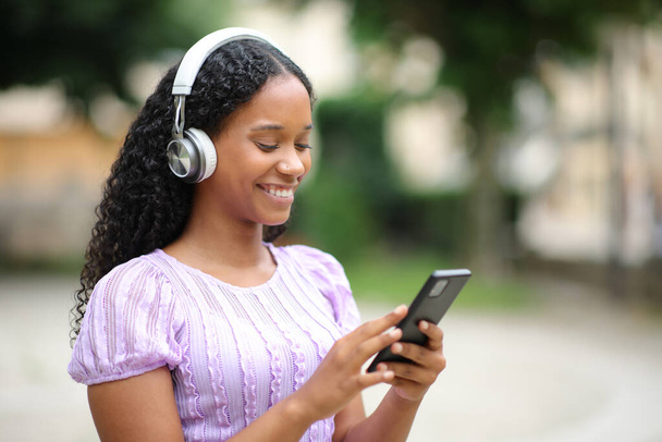 Mujer negra feliz con audífonos haciendo música en el teléfono caminando en la calle - Foto, imagen