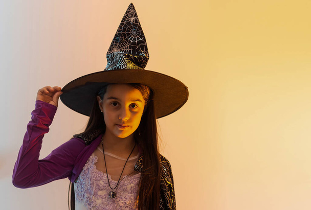 Menina adolescente bonito com cabelos longos morena em traje de bruxa preta, chapéu de pé perto da parede de madeira laranja. - Foto, Imagem