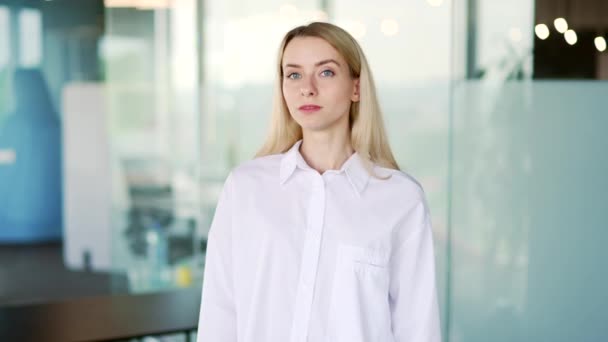 Portrait of a young smiling businesswoman standing in a modern office with crossed arms. Stylish happy blonde female employee in a white shirt is looking at the camera. Headshot of a successful manager - Video