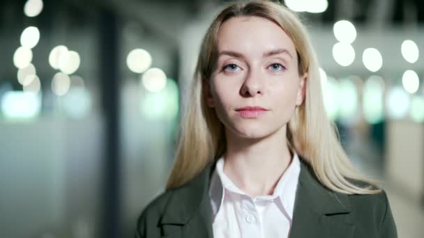 Close up. Portrait of a young smiling businesswoman standing in a modern office. Stylish happy blonde female employee in a suit looks at the camera. Headshot of a successful manager or entrepreneur - Filmagem, Vídeo