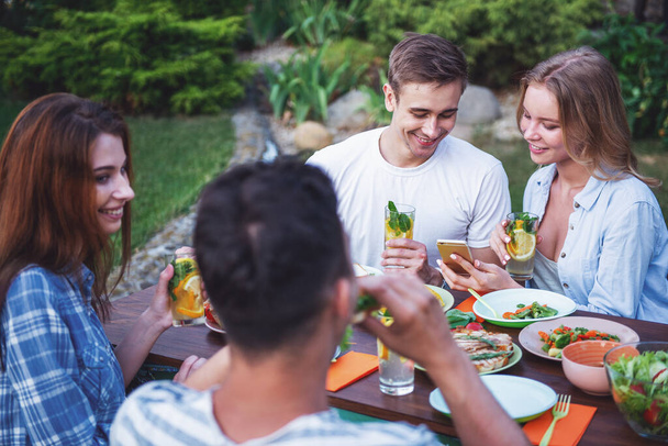 Young beautiful people are talking, using a smartphone, holding glasses and smiling while sitting at the table and having picnic outdoors - Photo, Image