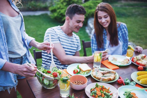 Young people are having picnic outdoors. Beautiful girl is mixing salad while her friends are talking, using a smartphone and smiling - Photo, Image