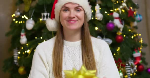 Nő télapó kalap ad karácsonyi ajándék doboz a kamera karácsonyfa háttér villogó fények koszorú. Boldog Új Évet, Boldog Karácsonyt. Kiváló minőségű 4k felvételek - Felvétel, videó