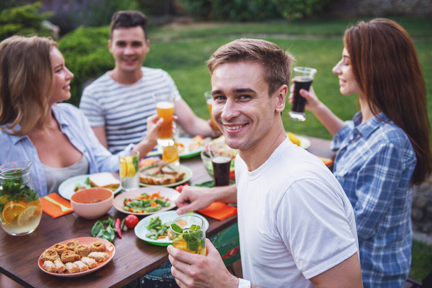 Mladí lidé jsou s piknik venku. Pohledný chlap drží sklenici limonády, při pohledu na fotoaparát a s úsměvem, zatímco jeho přátelé mluví v pozadí - Fotografie, Obrázek