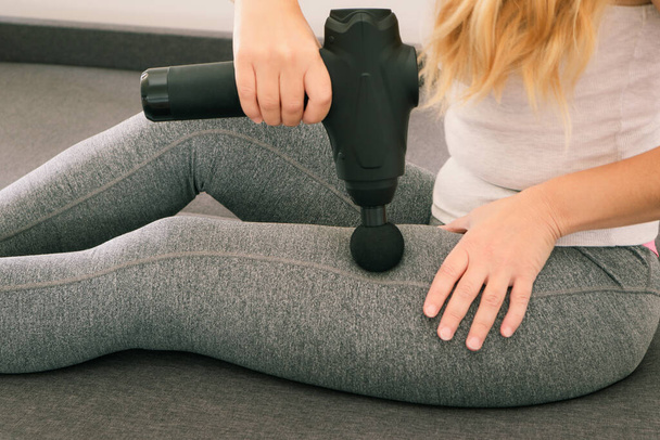 Selbstmassage der Frauenbeine mit einer Perfektionierungspistole zu Hause. Sportliche Frau mit therapeutisch perkussiver Massagepistole sitzt zu Hause auf dem Sofa. Eine Frau massiert ihre Beine mit einer automatischen Pistole - Foto, Bild