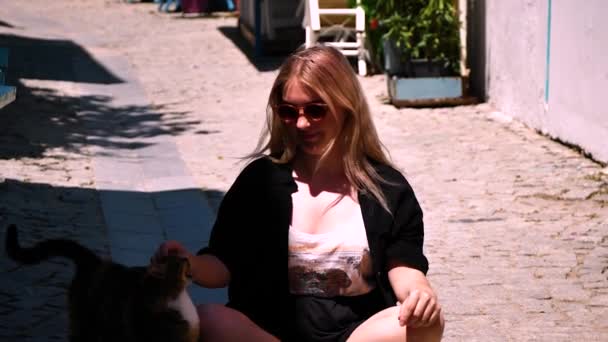 młoda blondynka dziewczyna w czarnym stroju kąpielowym w okularach przeciwsłonecznych i kapeluszu siedzi na molo na brzegu stret - Materiał filmowy, wideo