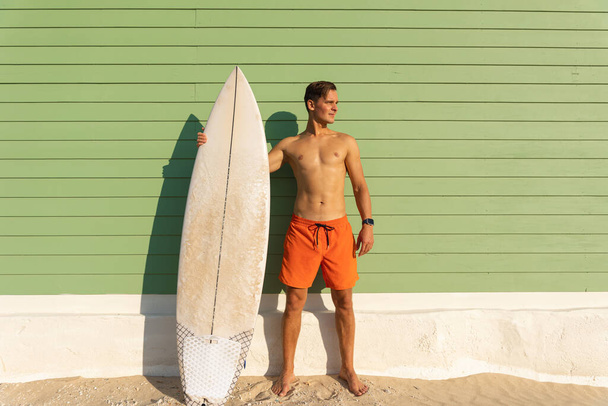 Un hombre con un bonito cuerpo de pie en la pared verde claro sosteniendo una tabla de surf mirando hacia un lado. Mid shot - Foto, Imagen