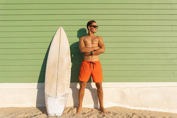 Ein lächelnder Mann mit schönem Körper mit Sonnenbrille steht am hellgrünen Rand mit einem Surfbrett - und schaut zur Seite. Mittelschuss - Foto, Bild