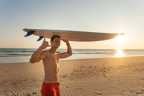 Привабливий усміхнений чоловік з гарним спортивним тілом тримає дошку для серфінгу однією рукою і дивиться в камеру, показуючи шаку. Середній постріл - Фото, зображення